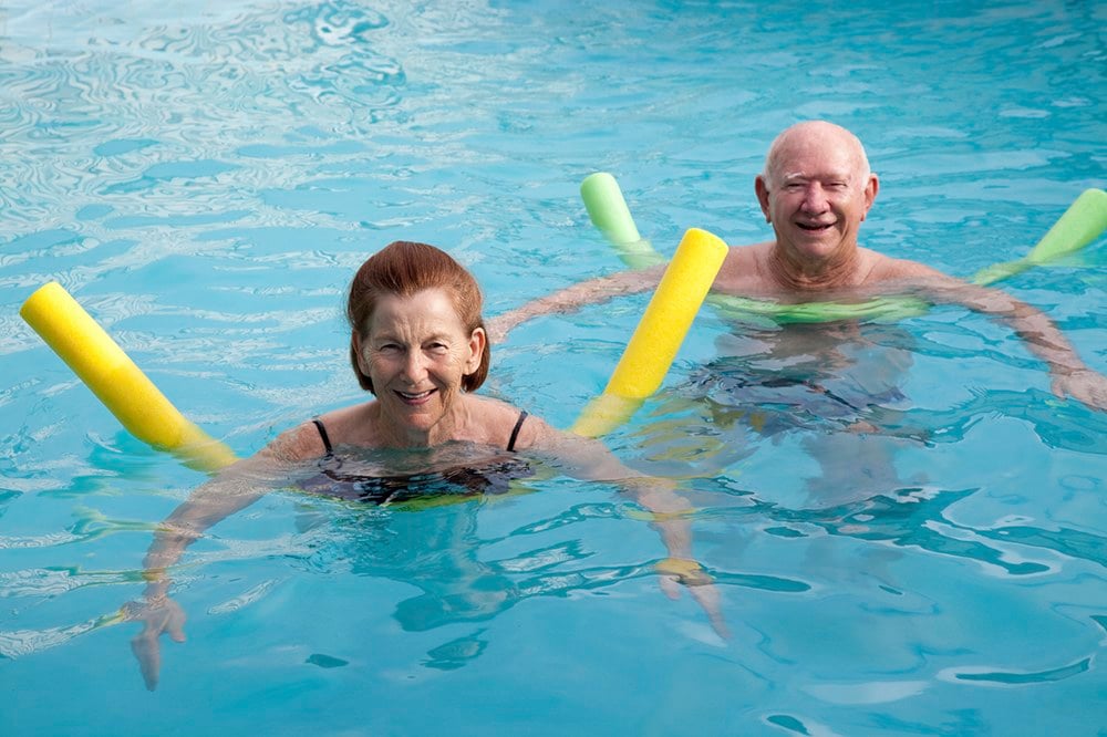 Kuvassa uima-altaassa vesinuudelien kanssa uivat iäkkäät nainen ja mies katsovat hymyillen kameraan.