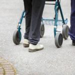 Kuvassa näkyvät ulkona kävelyllä olevan rollaattoria käyttävän vanhuksen jalat ja avustavan henkilön jalat.