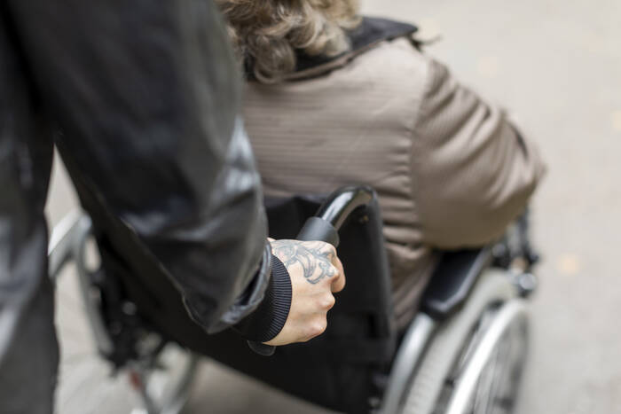 Kuvassa näkyy takaa kuvattuna henkilön käsi, joka auttaa ulkoilussa pyörätuolissa istuvaa henkilöä.