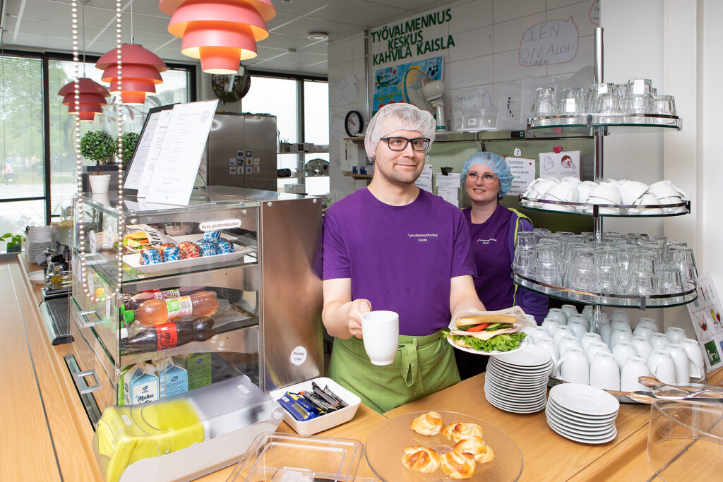 Kahvila Kaislan myyntitiski, jonka takaa miestyöntekijä ojentaa kahvikuppia ja lautasta, jolla on sämpylä. Ohjaaja seisoo taustalla.