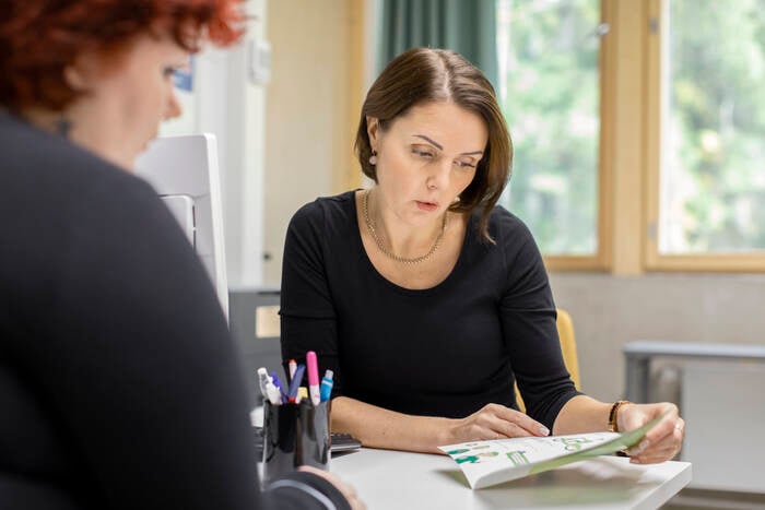 Tummahiuksinen nainen esittelee istuen pöydän ääressä esitettä punahiuksiselle naiselle.