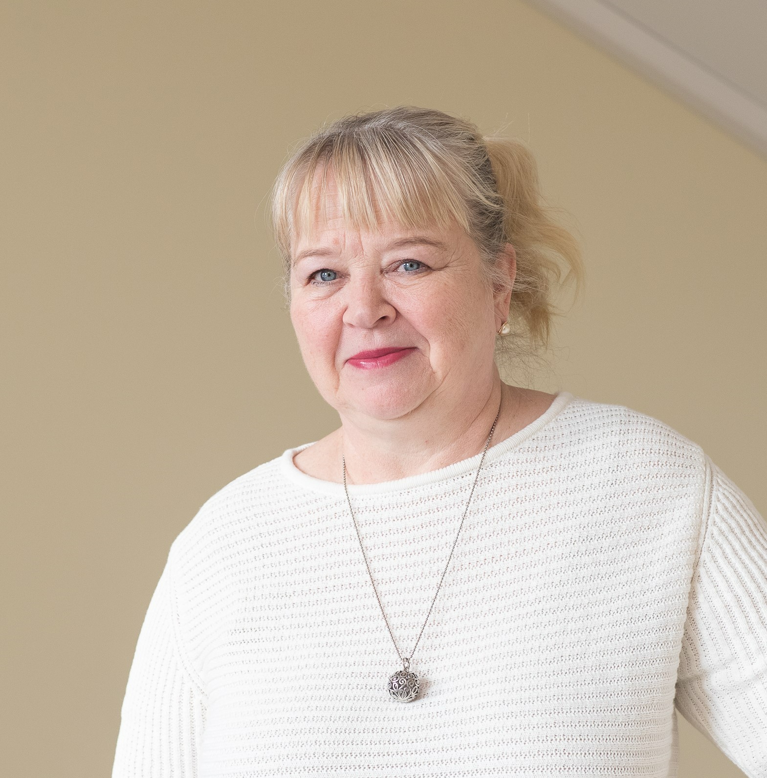 Kuvassa vaaleahiuksinen nainen, jolla päällään valkoinen neule ja kaulassaan riippuva kaulakoru