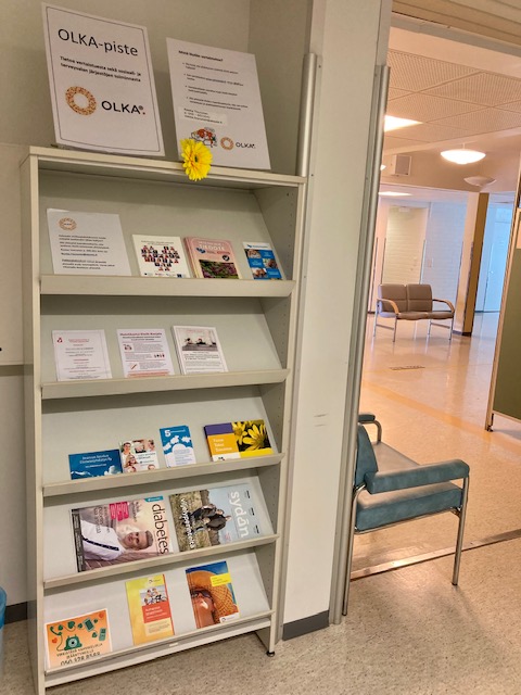 Hyvinvointiaseman käytävällä oleva hylly, jossa on järjestöjen esitteitä ja kyltti OLKA-piste.