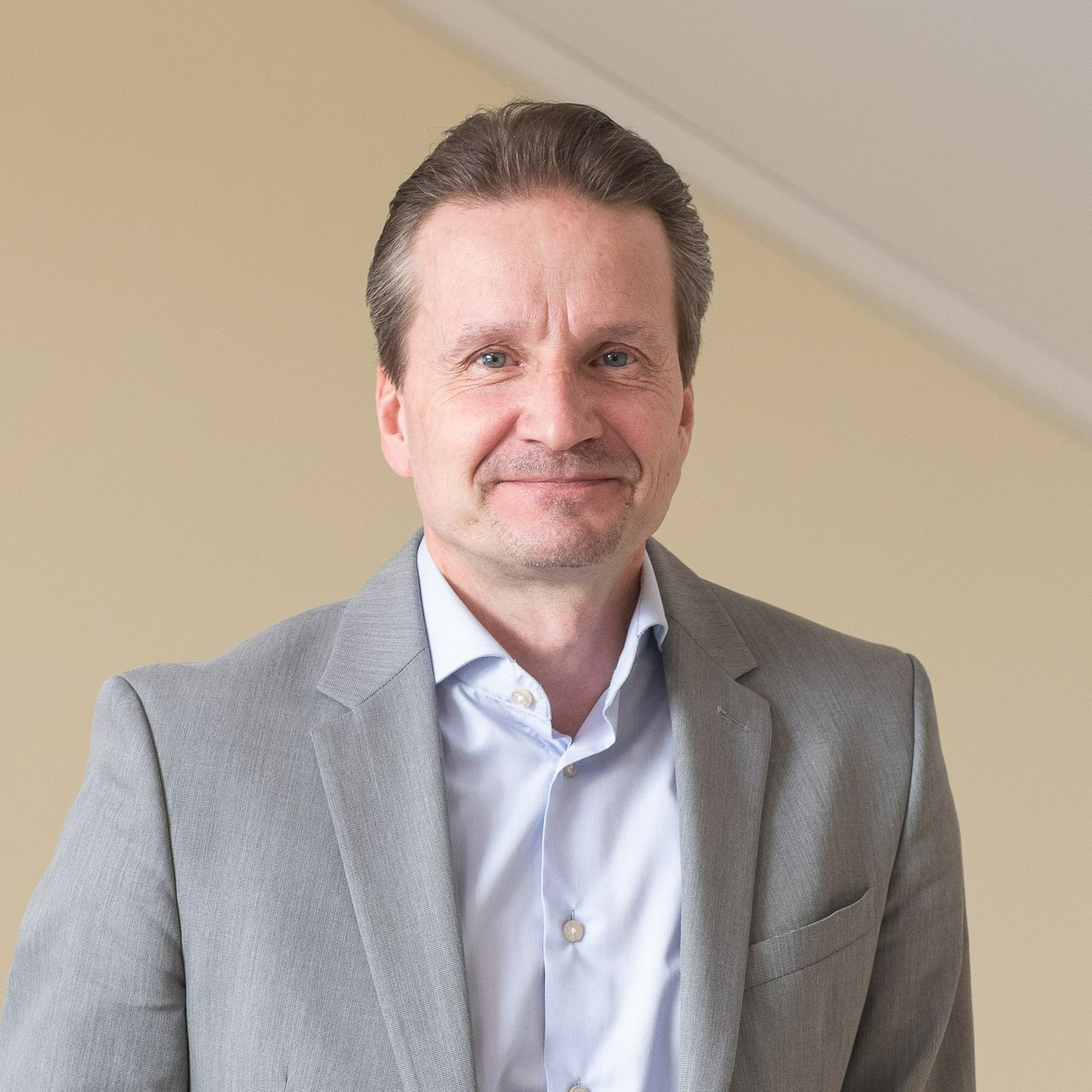 Valokuvassa tummahiuksinen hymyilevä mies, jolla valkoinen kauluspaita ja harmaa pikkutakki.