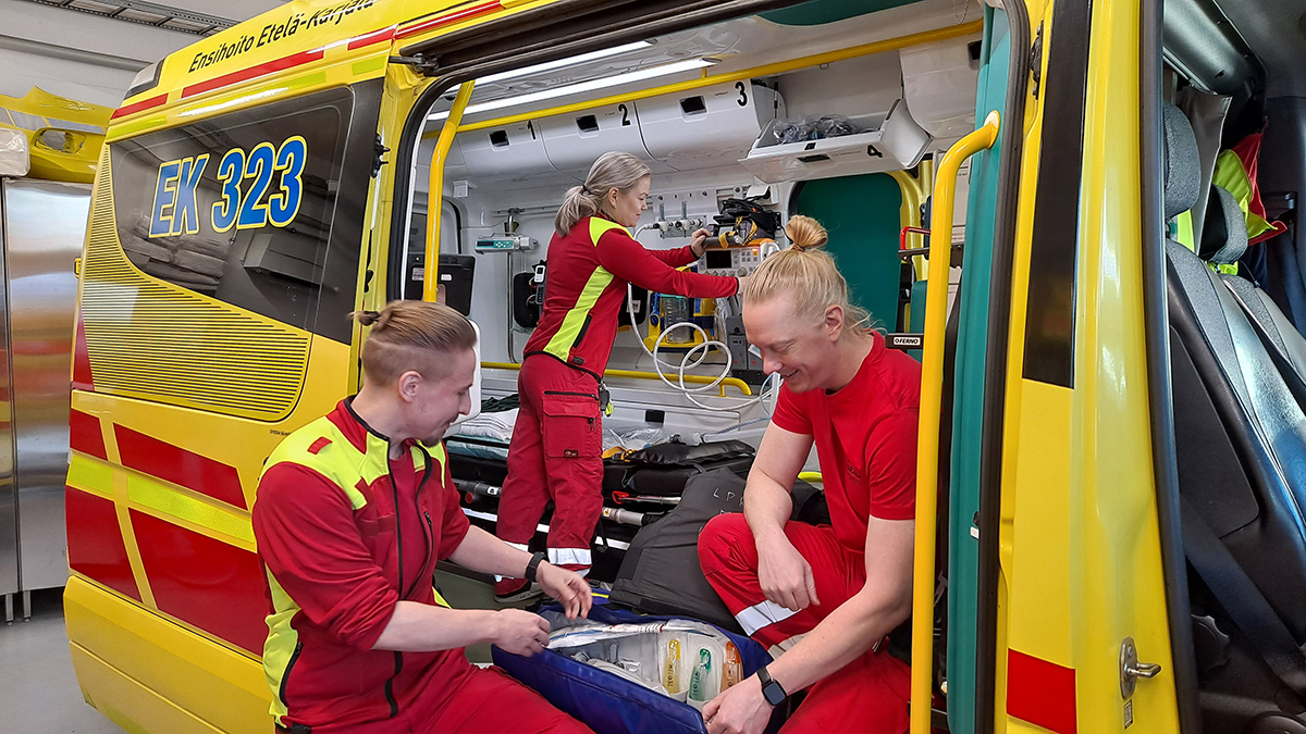 Ambulanssin avoimen oven edessä kyykistyneenä kaksi ensihoitajaa tutkimassa varustelaukun sisältöä ja yksi ensihoitaja järjestelee varusteita ambulanssin sisällä.