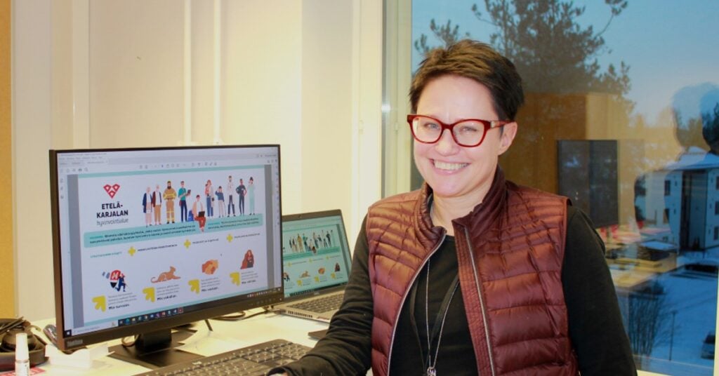 Tummahiuksinen punasankaiset silmälasit omaava nainen hymyilee työpöytänsä ääressä, taustalla tietokoneen näyttö ja ikkunasta avautuva talvinen maisema, päällään naisella on tummanpunainen toppaliivi ja musta paita.