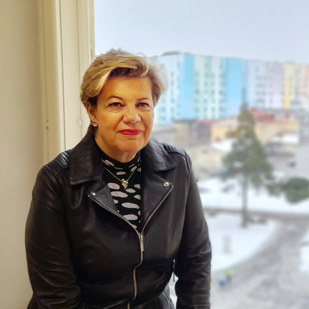 Vaalea lyhythiuksinen punahuulinen hymyilevä nainen istuu ikkunalaudalla ikkunan edessä ja ikkunasta avautuu taustalle talvinen näkymä Etelä-Karjalan keskussairaalan värikkääseen rakennukseen.