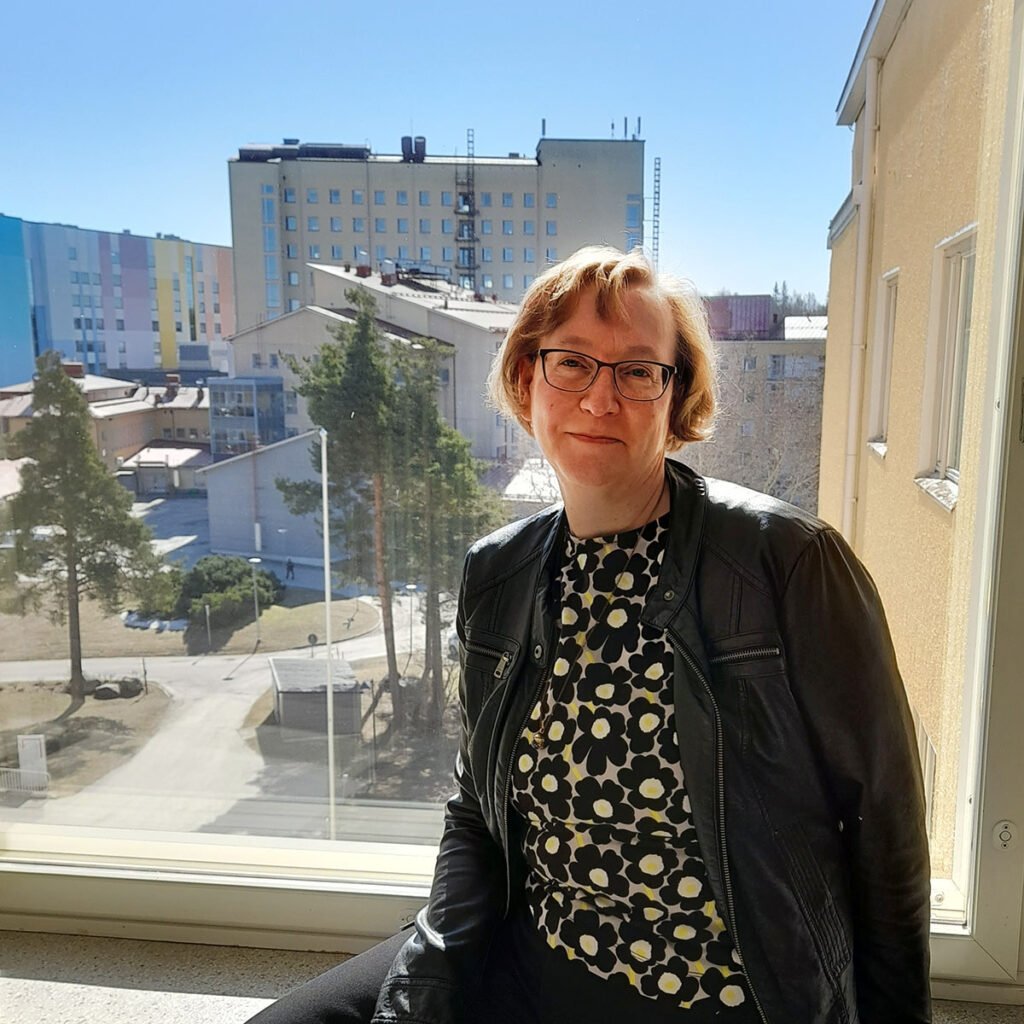 Hymyilevä vaalea lyhythiuksinen silmälasit silmillään oleva nainen istuu ikkunasyvennyksessä, ikkunasta avautuu aurinkoinen näköälä Etelä-Karjalan keskussairaalaan. 
