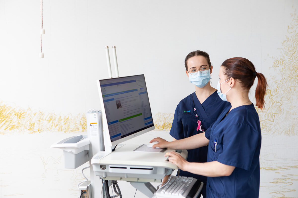 Tietokoneen ääressä seisomassa ja keskustelemassa kaksi sairaanhoitajanaista maskit kasvoillaan ja tummansiniset hoitaja-asut päällään.