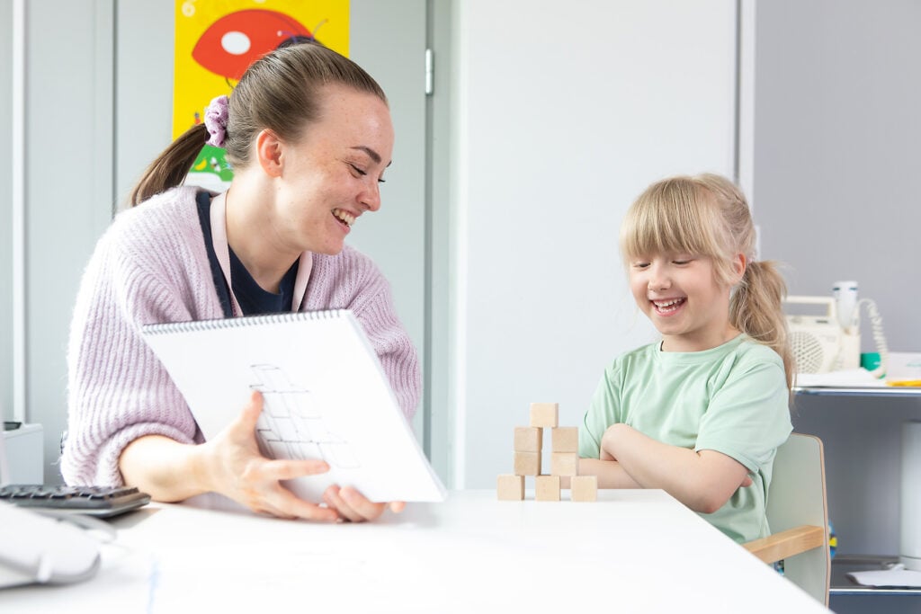 Neuvolassa hymyilevä hoitaja ja hymyilevä leikki-ikäinen lapsi katsovat yhdessä pöydälle rakennettua palikkarakennelmaa. Kuvassa on pastellinen värimaailma.