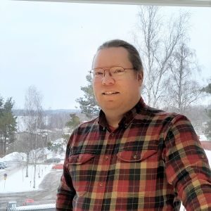 Hymyilevä silmälasipäinen mies, jolla päällään kirjavaruudullinen flanellipaita. Taustalla avautuu ikkunasta talvinen maisema.