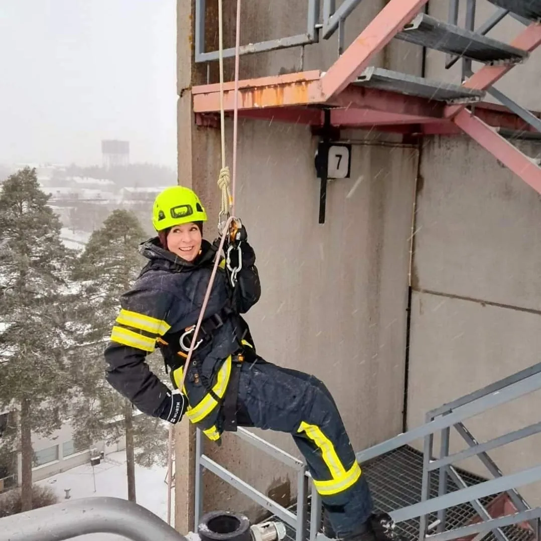 Hymyilevä nainen pelastajan varusteissa laskeutuu köyden varassa rakennuksen seinää pitkin alas.