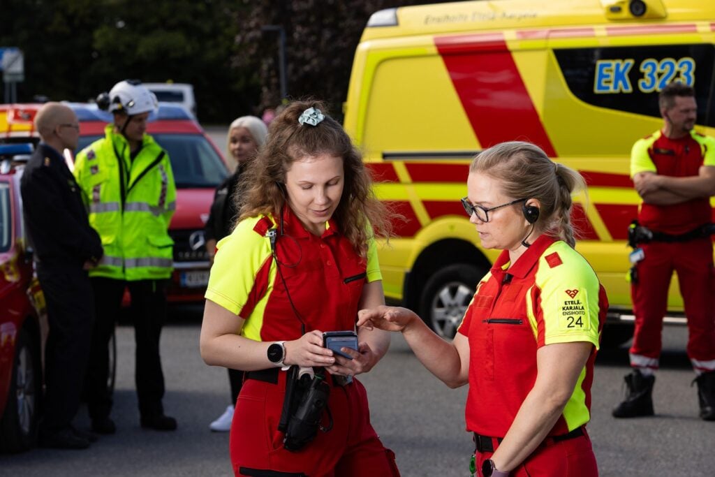 Tilannekuva, jossa etualalla kaksi ensihoitajan asussa olevaa naista tutkii kännykältä vaikuttavaa laitetta ja taustalla näkyy muita ihmisiä ja ambulanssi