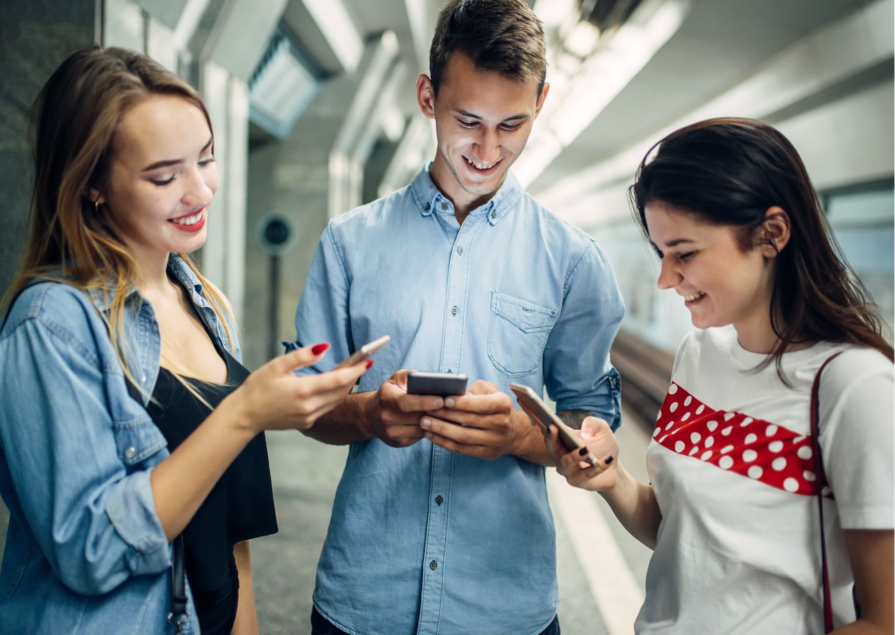 Kuvassa kolme nuorta (kaksi tyttöä ja yksi poika) seisovat ryhmässä ja katsovat kännyköitään iloisina