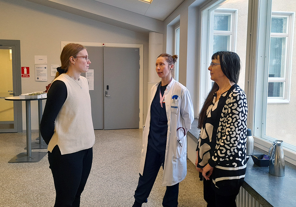 Kuvassa kolme naista keskustelee käytävällä keskenään toisiinsa katsoen, keskimmäisellä naisella on lääkärintakki yllään.