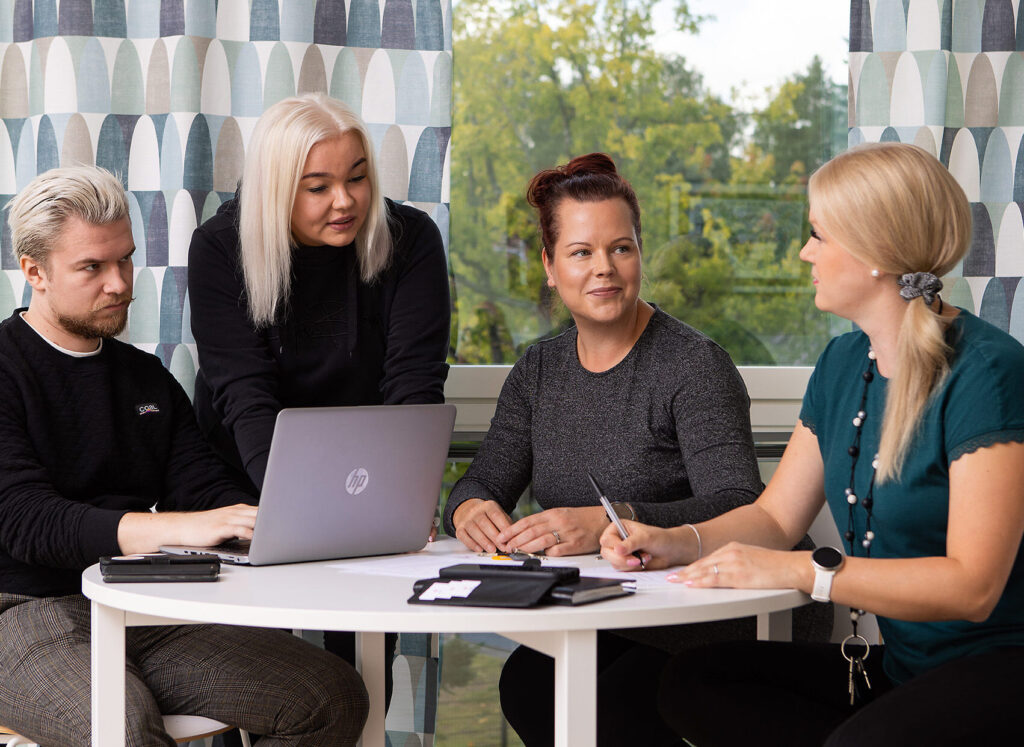 Kuvassa neljä ihmistä istuu pöydän ääressä keskustelemassa. Yhdellä on tietokone ja yksi kirjaa asioita paperille. Yhdellä on avaimet kaulassaan. Henkilöt ovat pukeutuneet tavallisiin siviilivaatteisiin.