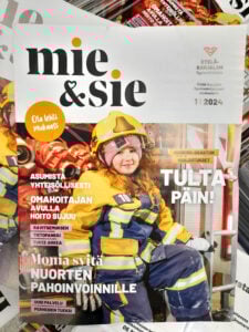 Kuvassa Mie&Sie-lehden kansikuva, jossa palomiehen pukuun pukeutunut hymyilevä tyttö istuu paloauton oviaukossa ja katsoo kameraan. Kuvassa on paljon oranssia, keltaista ja tummansinistä väriä.