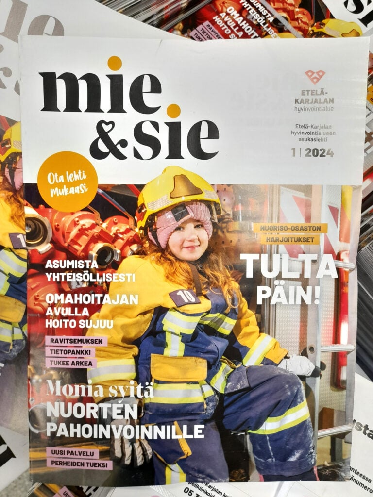 Kuvassa Mie&Sie-lehden kansikuva, jossa palomiehen pukuun pukeutunut hymyilevä tyttö istuu paloauton oviaukossa ja katsoo kameraan. Kuvassa on paljon oranssia, keltaista ja tummansinistä väriä. 
