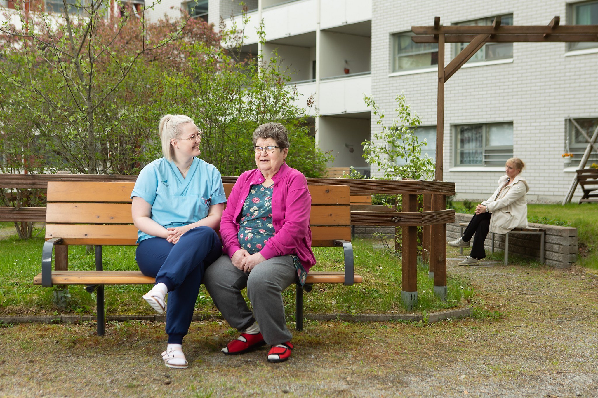 Työntekijä ja iäkäs nainen istuvat puiston penkillä kääntyneenä toisiaan kohden ja näyttävät iloisilta sekä rauhallisilta