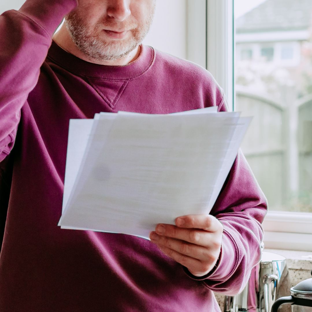 Lähikuva viininpunaiseen collegepuseroon pukeutuneesta miehestä, josta ei näy kuin rintakehä ja hieman parrakasta leukaa. Mies pitää valkoista paperinippua käsissään.