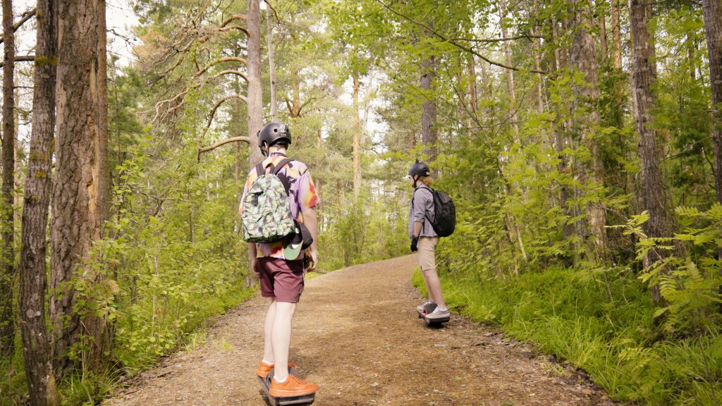 Kaksi nuorta onewheelaa selät kameraan päin hiekkatietä metsässä. Nuorilla on kypärät päässä.