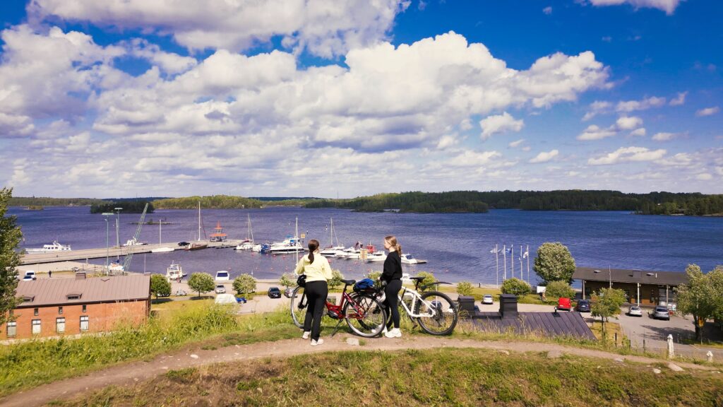Kaksi pyöräilee seisoo kesäisessä kuvassa pyöriensä kanssa Lappeenrannan valleilla kulkevalla polulla. Taustalla näkyy Lappeenrannan satamaa ja Saimaata.