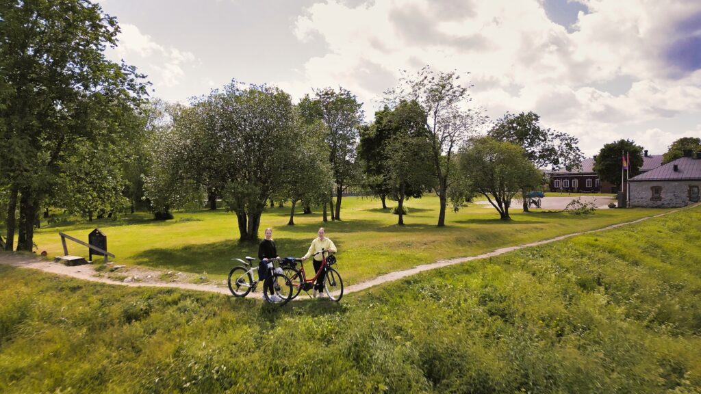 Kaksi pyöräilee seisoo kesäisessä kuvassa pyöriensä kanssa Lappeenrannan valleilla kulkevalla polulla. Taustalla näkyy linnoituksen puistoa.