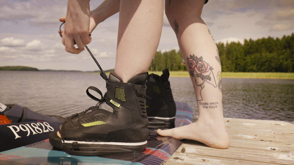 Kesäisessä kuvassa paljasjalkainen henkilö solmii wakeboardia jalkoihinsa. Henkilö seisoo laiturilla, taustalla näkyy järvi.