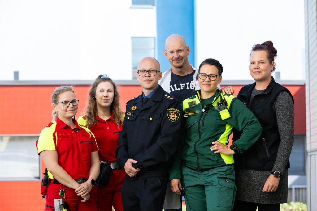 Ryhmäkuva kuudesta Etelä-Karjalan hyvinvointialueen työntekijästä eri ammateissa. Etualalla ensihoitajia punaisissa ja vihreissä asuissa sekä virkapukuinen pelastusalan työntekijä. Taustalla kaksi siviilivaatteissa olevaa henkilöä. Kuva on otettu ulkona sairaalarakennuksen edustalla.