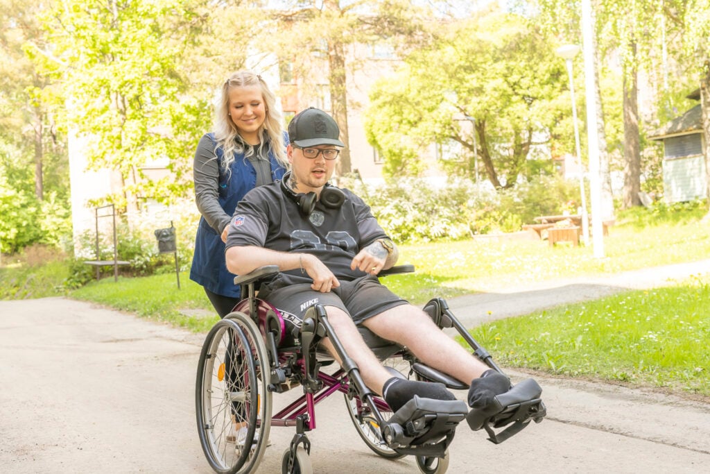 Lähihoitaja hymyilee ja työntää asiakasta (nuori mies) pyörätuolissa