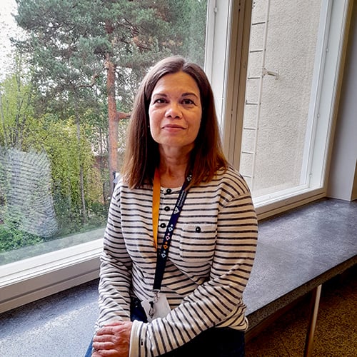 Hymyilevä ruskeatukkainen nainen katsoo kameraan pieni hymynkare huulillaan. Hän istuu leveällä ikkunalaudalla ja taustalla avautuu metsäinen maisema. Hänellä on päällään valko-musta-raidallinen pitkähihainen paita ja farkut. Kuva on rajattu näyttämään hänet lantiosta ylöspäin.