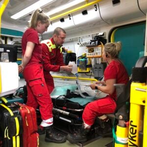 Ensihoidon lehtori, ensihoitajaopiskelija ja kehittäjäensihoitaja ambulanssin takatilassa opettelemassa potilaan hoidossa tarvittavia taitoja.