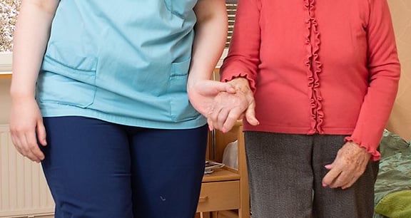 Sinisiin pukeutunut ihminen taluttaa punaisessa paidassa olevaa ikäihmistä kädestä. Kuvassa näkyy keskivartalot.