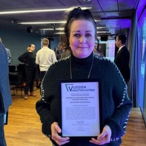 Kuvassa Kati Saarikivi pitää palkintotaulua. Takan muita ihmisiä juhlassa.