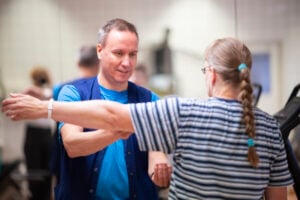 Kasvot kameraan päin oleva fysioterapeuttimies ohjaa selkä kameraan olevaa naishenkilöä. Naisella on käsi ojennettuna suoraan sivulla ja fysioterapeutti ohjaa käden liikettä.