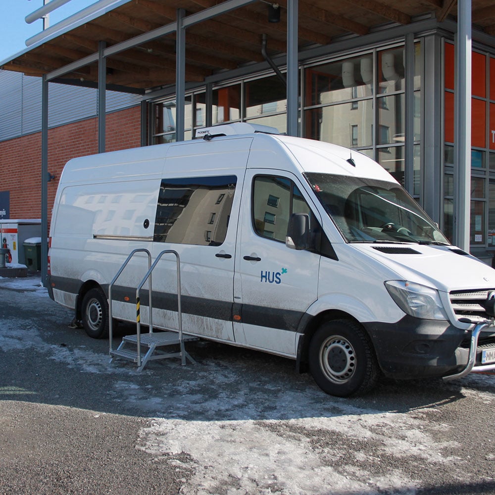 Vinottain kuvassa näkyy valkoinen kookas pakettiauto, jossa etuovessa teksti HUS. Sivuoven edessä on siirrettävä metallinen porrasaskelma, jossa korkeat kaiteet. Taustalla näkyy punatiilinen liikerakennus. Valokuvasta välittyy auringonpaiste ja hiekkaisessa maassa on hieman valkoista lunta.