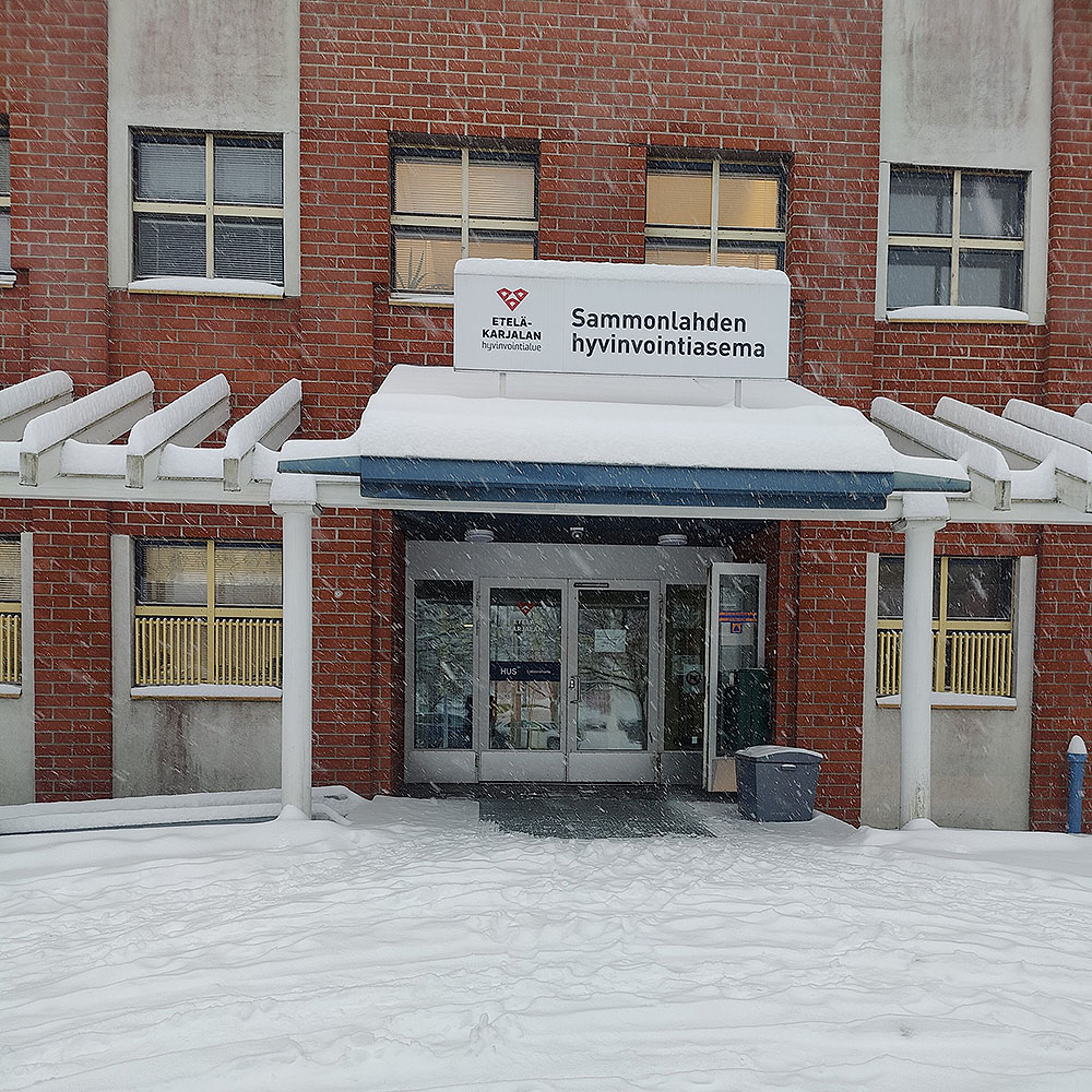 Punatiilinen rakennus lumisateessa. Rakennuksen sisäänkäynnillä on sininen katos, ja sen yläpuolella on kyltti, jossa lukee 'Sammonlahden hyvinvointiasema' sekä Etelä-Karjalan hyvinvointialueen logo. Maa ja katto ovat lumen peitossa, ja ovien edessä näkyy poljettu lumipinta.