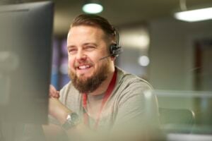 Mies istuu luurit päässä tietokoneen edessä ja hymyilee luurit päässä