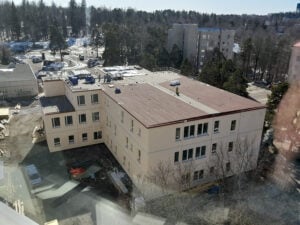 Ilmakuva rakennustyömaasta, jossa kootaan moduulirakenteista rakennusta. Rakennus on kolmikerroksinen, vaaleasävyinen ja sen katolla on laitteita sekä rakennusmateriaaleja. Rakennuksen ympärillä on edelleen työmaa, mutta rakennus on lähes valmis. Taustalla näkyy puita, kerrostaloja ja lumisia alueita.