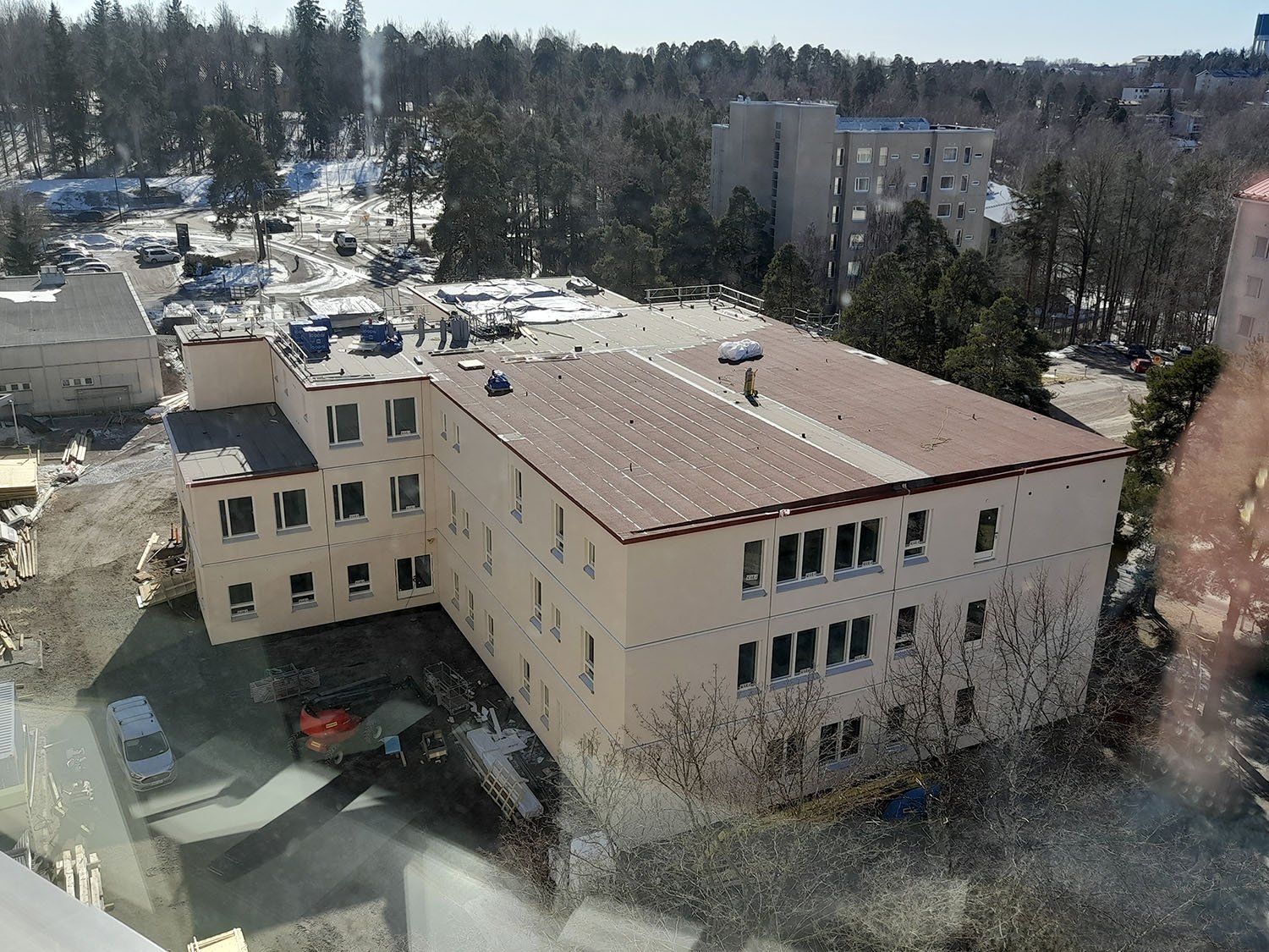 Ilmakuva rakennustyömaasta, jossa kootaan moduulirakenteista rakennusta. Rakennus on kolmikerroksinen, vaaleasävyinen ja sen katolla on laitteita sekä rakennusmateriaaleja. Rakennuksen ympärillä on edelleen työmaa, mutta rakennus on lähes valmis. Taustalla näkyy puita, kerrostaloja ja lumisia alueita.