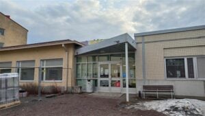 Kuva esittää matalaa rakennusta, jossa on lasiovinen sisäänkäynti. Oven päällä on harjakattoinen katos, ja oven vieressä on punainen "F"-kirjain. Rakennuksen vasemmalla puolella on keltaiseksi maalattu osa, jossa on useita ikkunoita. Oikealla puolella on vaaleankeltainen tiiliseinä, jossa on myös ikkunoita. Edessä on sorapintainen piha, jossa on hieman lunta ja rakennusmateriaaleja sekä metallinen aita. Taustalla näkyy osittain pilvinen taivas.