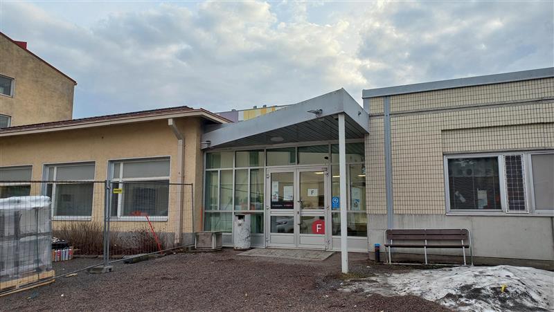 Kuva esittää matalaa rakennusta, jossa on lasiovinen sisäänkäynti. Oven päällä on harjakattoinen katos, ja oven vieressä on punainen "F"-kirjain. Rakennuksen vasemmalla puolella on keltaiseksi maalattu osa, jossa on useita ikkunoita. Oikealla puolella on vaaleankeltainen tiiliseinä, jossa on myös ikkunoita. Edessä on sorapintainen piha, jossa on hieman lunta ja rakennusmateriaaleja sekä metallinen aita. Taustalla näkyy osittain pilvinen taivas.