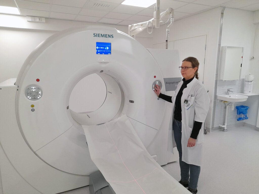 Hoitohenkilö seisoo Siemensin Biograph mCT -laitteen vieressä, valmiina käynnistämään kuvauksen. Kuvassa näkyy moderni sairaalaympäristö ja potilaspöytä, joka ulottuu laitteen tunnelin läpi.