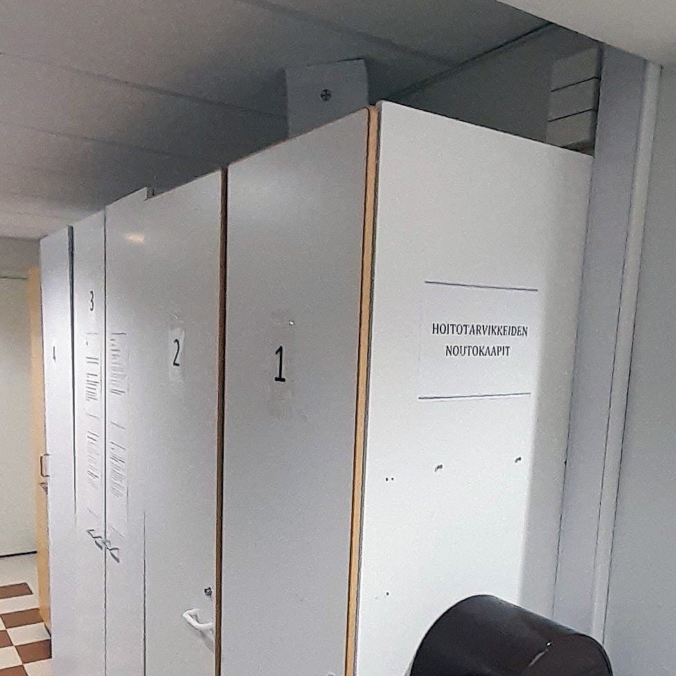 Rivi harmaita kaappeja, joiden ovet on numeroitu 1, 2 ja 3. Yhdessä kaapin ovessa on myös teksti "HOITOTARVIKKEIDEN NOUTOKAAPIT". Taustalla näkyy valkoinen käytävä ja ruudullinen lattia.