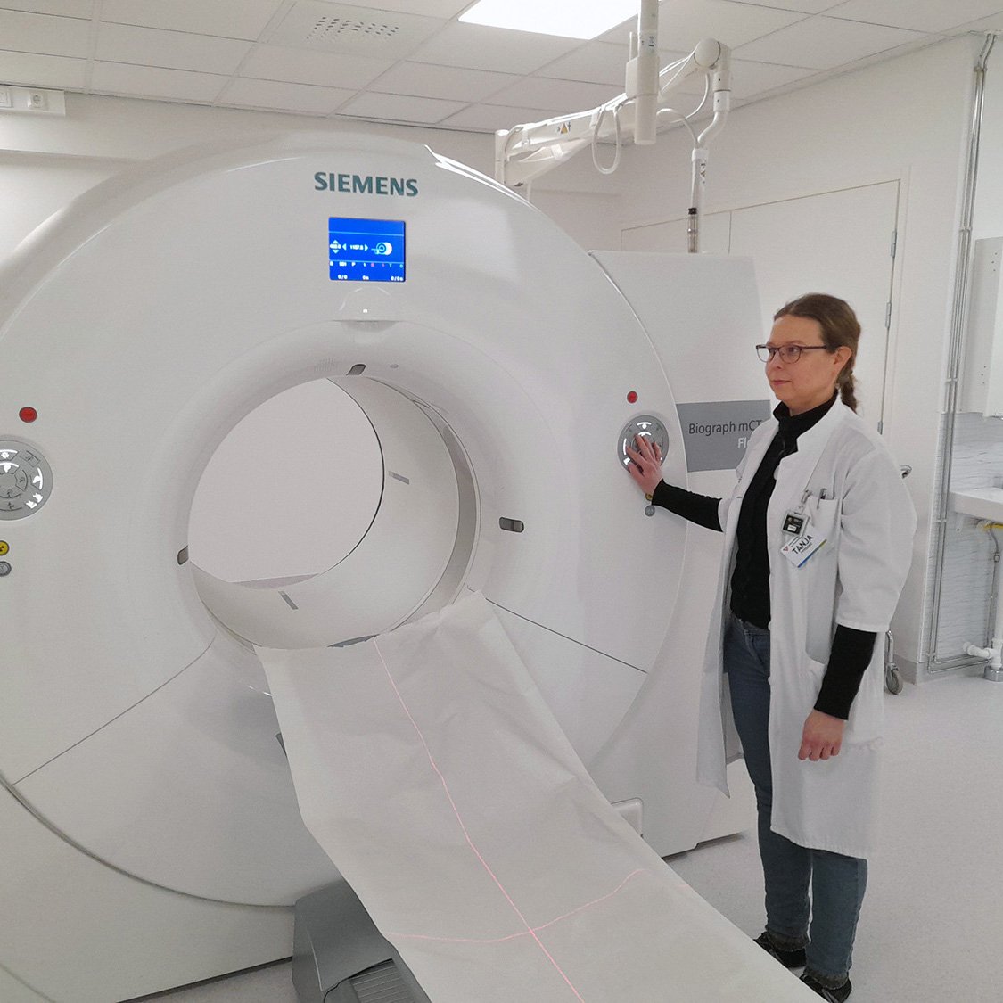 Hoitohenkilö seisoo Siemensin Biograph mCT -laitteen vieressä, valmiina käynnistämään kuvauksen. Kuvassa näkyy moderni sairaalaympäristö ja potilaspöytä, joka ulottuu laitteen tunnelin läpi.