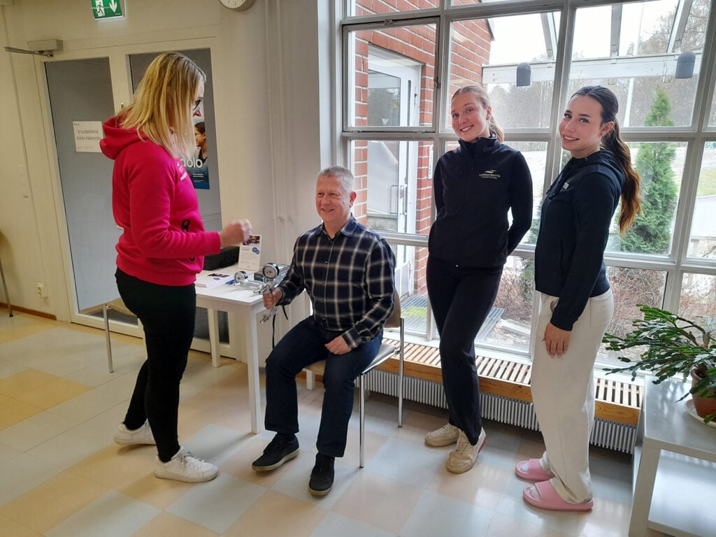 Nainen mittaa miehen puristusvoimaa kädessä pidettävällä mittarilla sisätiloissa. Kaksi muuta naista seisoo vieressä hymyillen. Taustalla suuri ikkuna, josta näkyy ulos. Tilanne vaikuttaa olevan osa terveyteen liittyvää tapahtumaa tai mittauspistettä.
