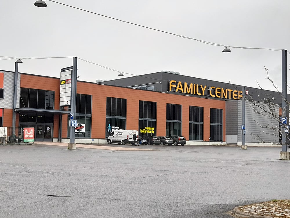 Kauppakeskus nimeltään "Family Center" harmaana ja pilvisenä päivänä. Rakennus on moderni, kaksikerroksinen ja julkisivultaan punatiilen ja harmaan sävyinen. Etualalla on iso, tyhjä parkkialue ja muutamia pysäköityjä autoja. Rakennuksessa näkyvät useiden yritysten kyltit, kuten Masku ja Saimaan Työtarvike Oy.