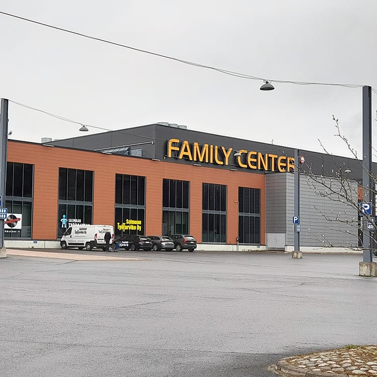 Kauppakeskus nimeltään "Family Center" harmaana ja pilvisenä päivänä. Rakennus on moderni, kaksikerroksinen ja julkisivultaan punatiilen ja harmaan sävyinen. Etualalla on iso, tyhjä parkkialue ja muutamia pysäköityjä autoja. Rakennuksessa näkyvät useiden yritysten kyltit, kuten Masku ja Saimaan Työtarvike Oy.