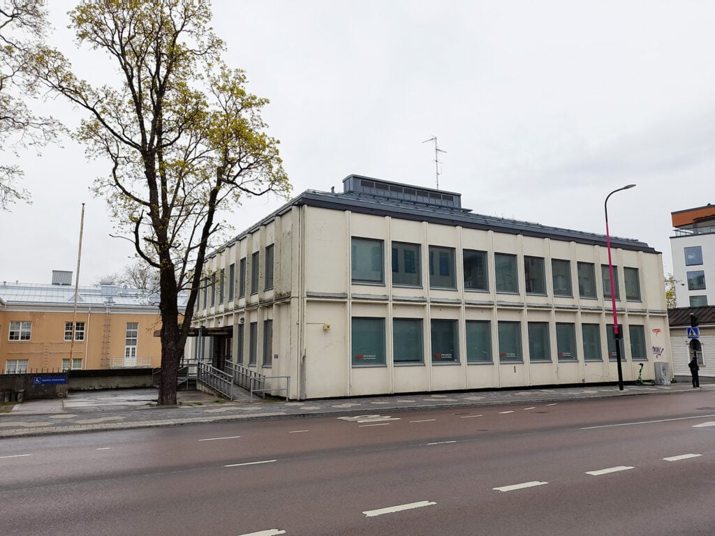 Vaalea kaksikerroksinen virastomainen rakennus kadun varrella harmaana ja sateisena päivänä. Rakennuksessa on useita samanlaisia ikkunoita, joissa on sälekaihtimet. Etualalla näkyy jalkakäytävä ja ajotie, jossa on valkoisia kaistaviivoja. Rakennuksen edessä on esteetön luiska, ja vasemmalla kasvaa iso lehtipuu. Taustalla näkyy lisää rakennuksia, muun muassa vaaleankeltainen ja moderni lasipintainen talo.