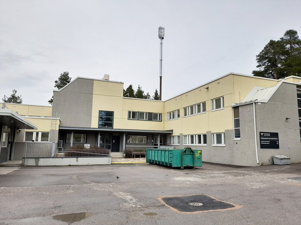 Kaksikerroksinen vaaleankeltainen ja harmaa julkinen rakennus, jonka edessä on asfalttipiha ja kaksi vihreää jäteastiaa. Rakennus on moderni mutta hillityn näköinen, ja sen julkisivussa on sekä tiiltä että sileää pintaa. Sisäänkäynnin yläpuolella on pieni katos. Taustalla näkyy metsää ja korkea matkapuhelinverkkomasto. Oikealla on musta opastekyltti, jossa lukee "Etelä-Karjalan hyvinvointialue". Sää on pilvinen ja harmaa.
